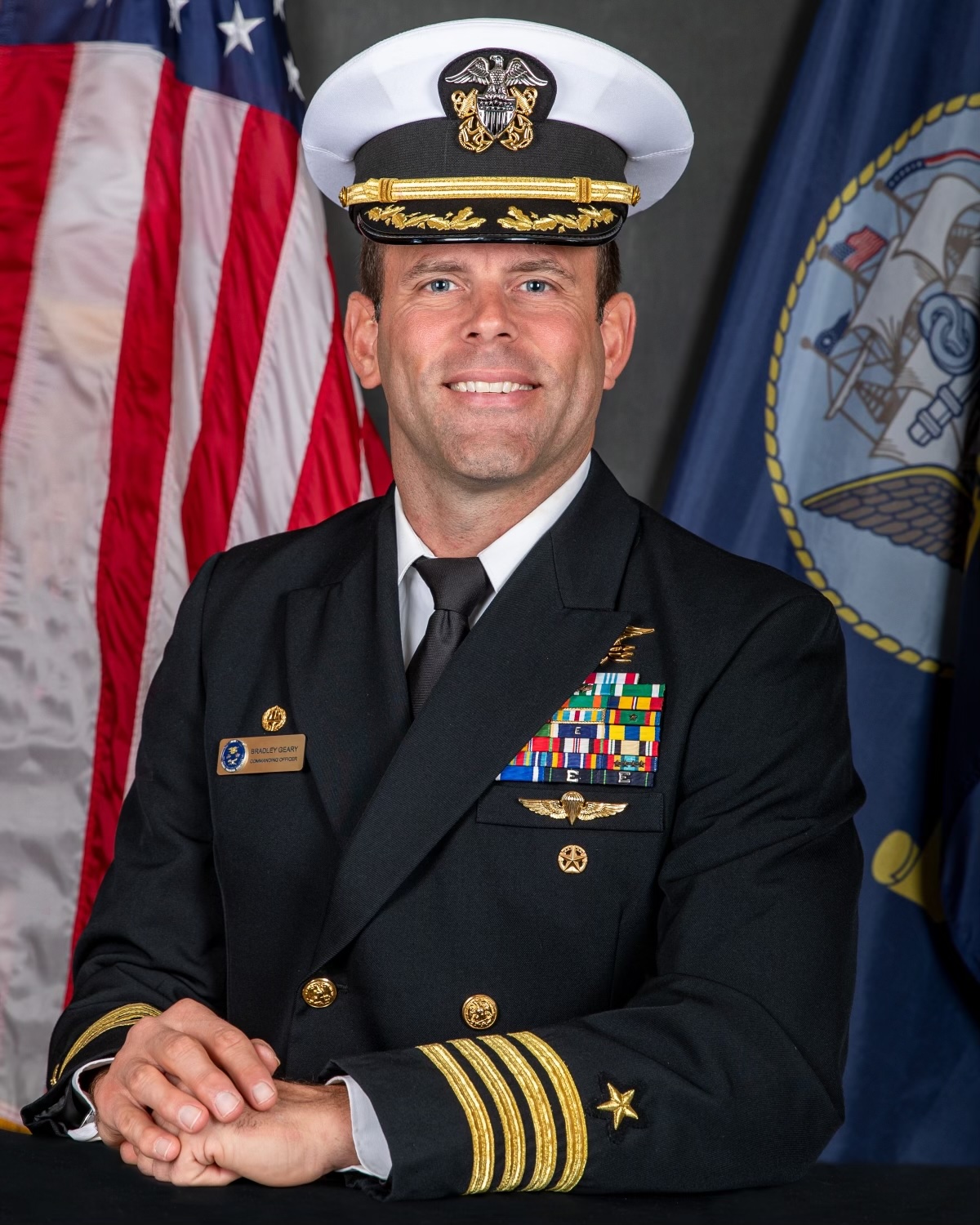 Portrait of Bradley Geary, a brown-haired man smiling in uniform, with U.S. and Navy flags in the background.