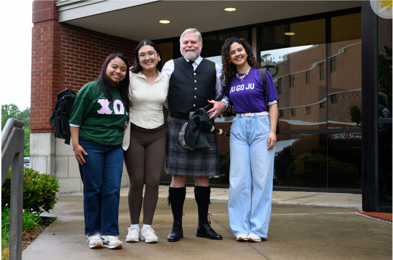 Three female students pose with a man with a white beard in a kilt and a black vest with gold buttons, holding a bagpipe