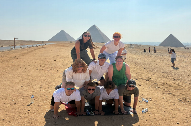 Eight students are arranged in a pyramid by kneeling on each other's backs, in front of pyramids in Egy[t