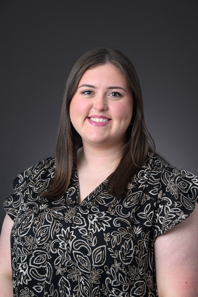 Rosalyn Cofer, a woman with brown hair wearing a black and gold patterned blouse smiling on a gray background.