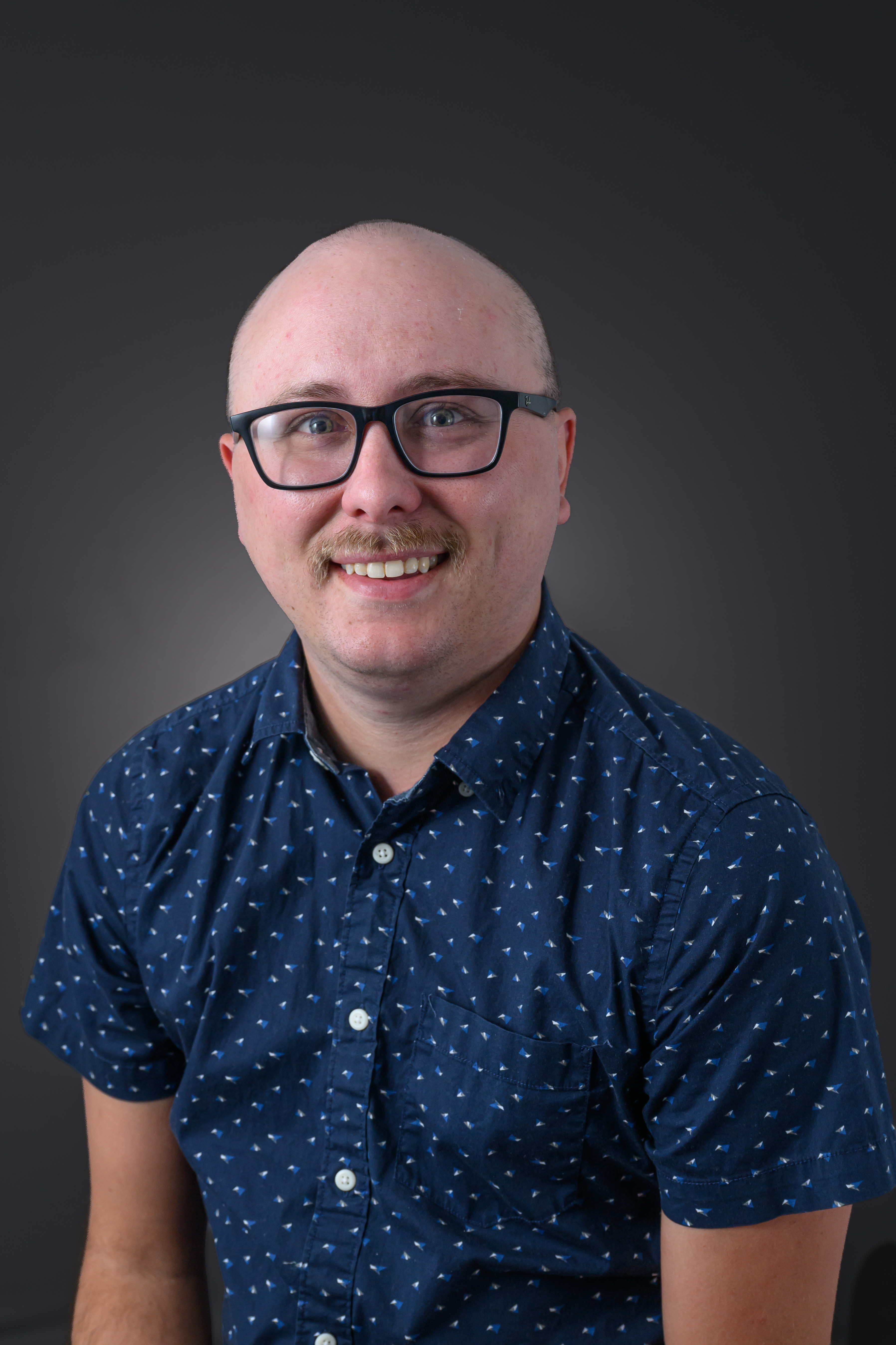 Taylor Hodges, a man smiling with glasses wearing a dark blue patterned collared shirt against a gray background.