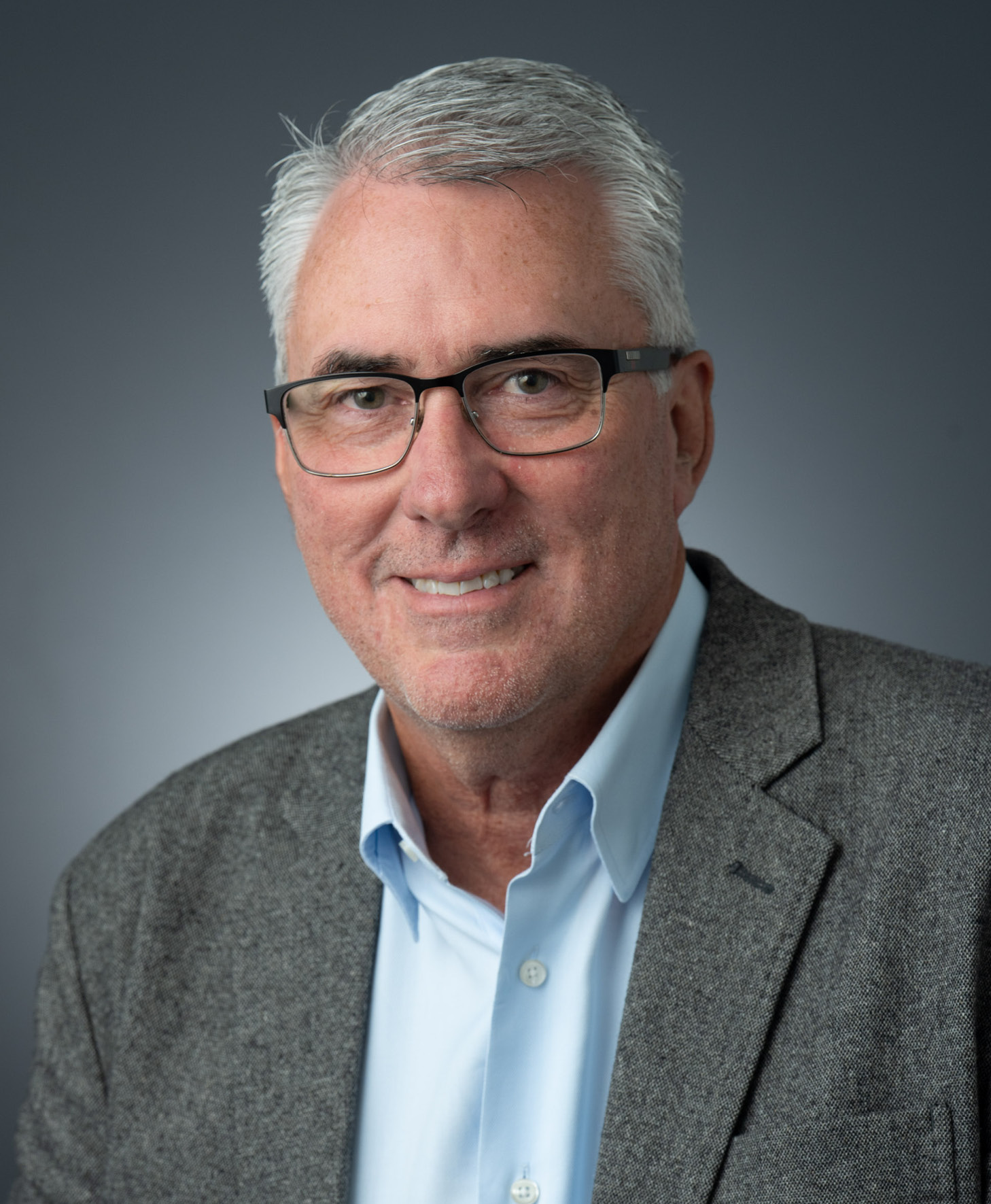 Todd McCullough, smiling man with gray hair and glasses, wearing gray blazer and blue dress shirt, against gray background.