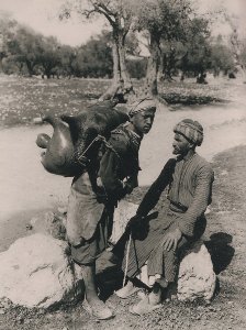 two men, one is sitting and one is standing wearing a water skin on his back