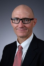 Professional headshot of Michael McGalliard Dean of the College of Allied Health and Associate Provost for Health Sciences. A bald man wearing glasses, a dark suit, a white shirt, and a red patterned tie, posed against a neutral gray background.