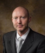 Headshot of Dr. Morten Jenson, a man wearing a dark suit and silver tie, against a brown studio background.