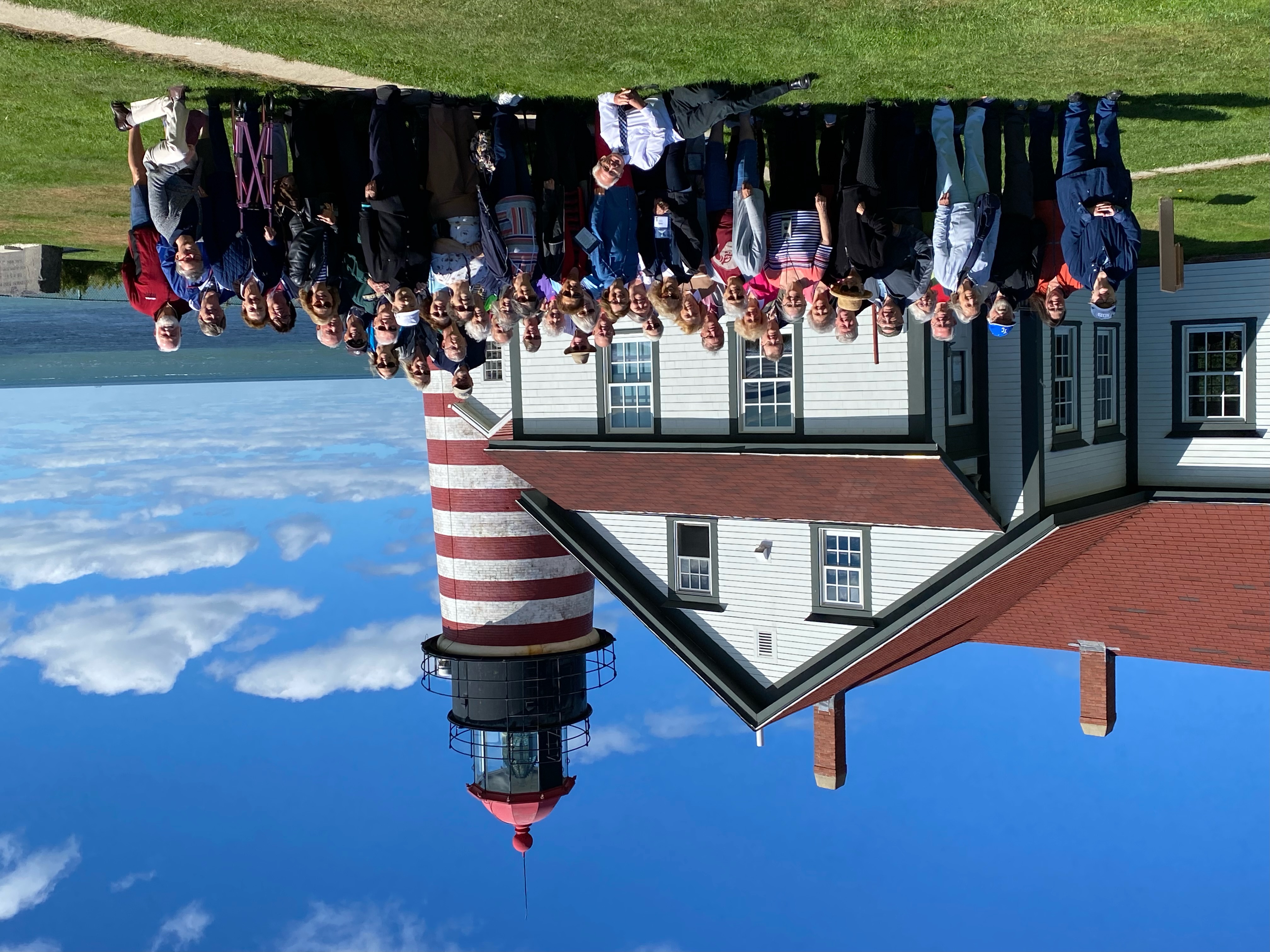 A large group of older adults posing together in front of a white house next to a red-and-white striped lighthouse, near the ocean under a bright blue sky.