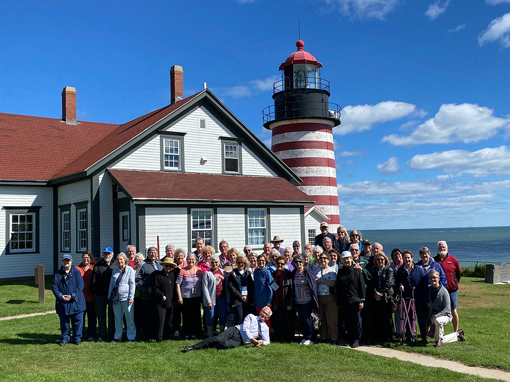 A large group of older adults posing together in front of a white house next to a red-and-white striped lighthouse, near the ocean under a bright blue sky.