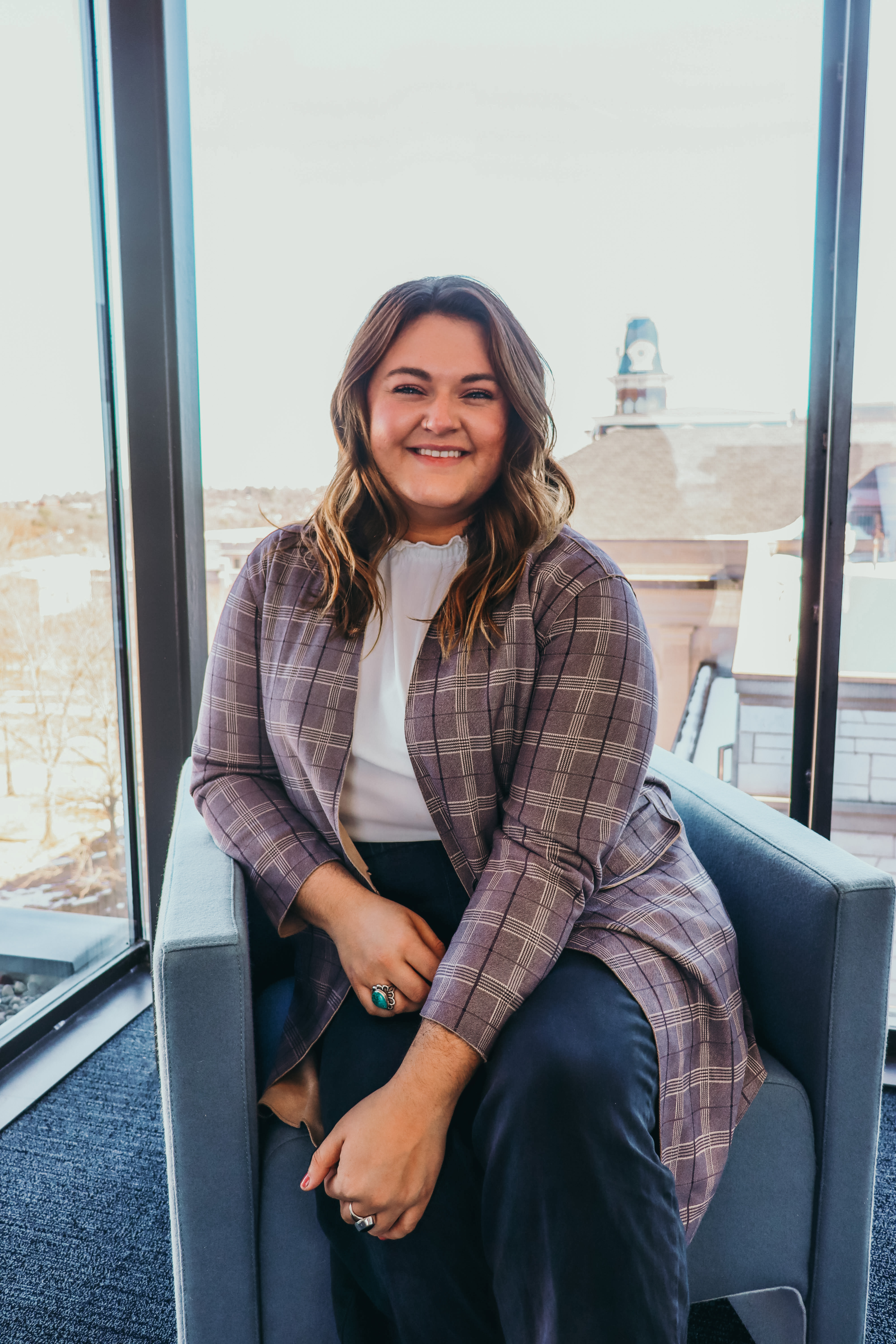 Shannon seated in a chair by a large window, wearing a plaid jacket and dark pants with a city view behind.