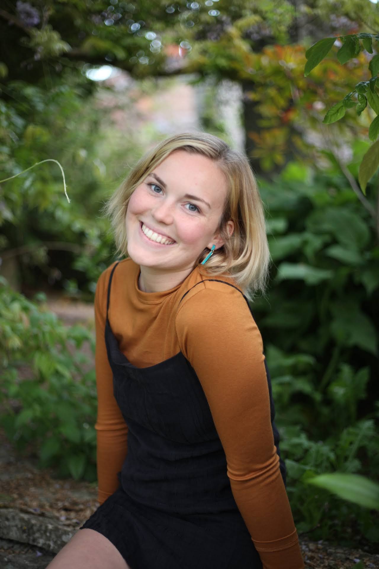 Jenna smiling outdoors, wearing a rust top and black dress, surrounded by greenery.