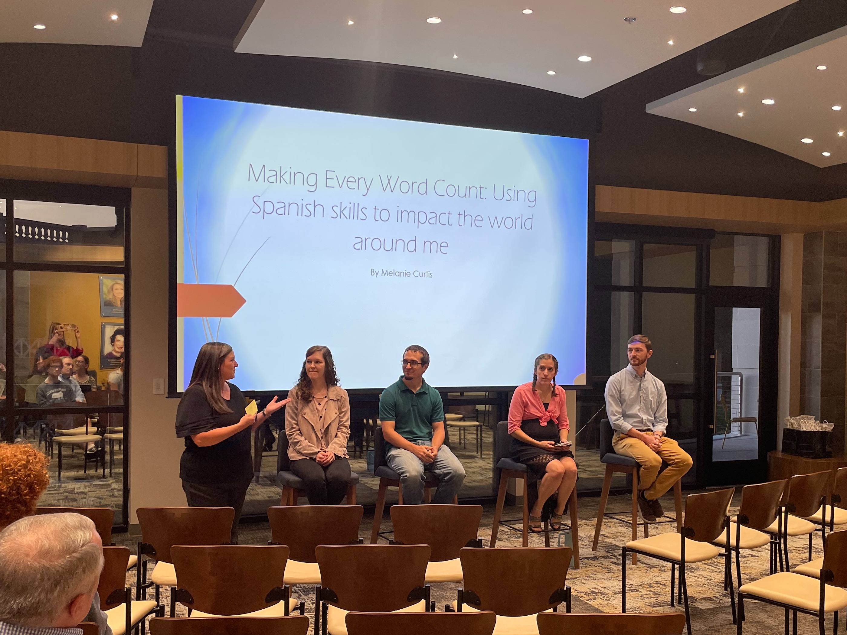 A panel of four students sit on stools in an auditorium with a powerpoint behind them that says, 'Making Every Word Count: Spanish skills to  impact the world around me'