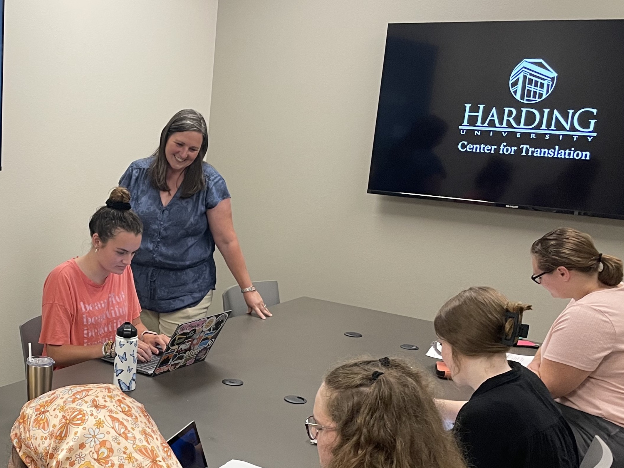A professor in a blue shirt looks over a group of students working on their laptops around a table