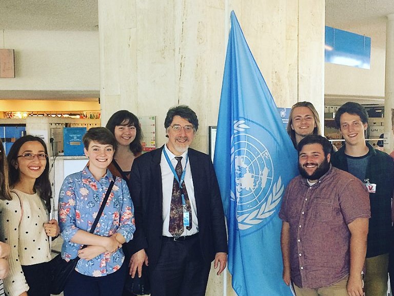 Six students are standing around a United Nations flag and a United Nations representative