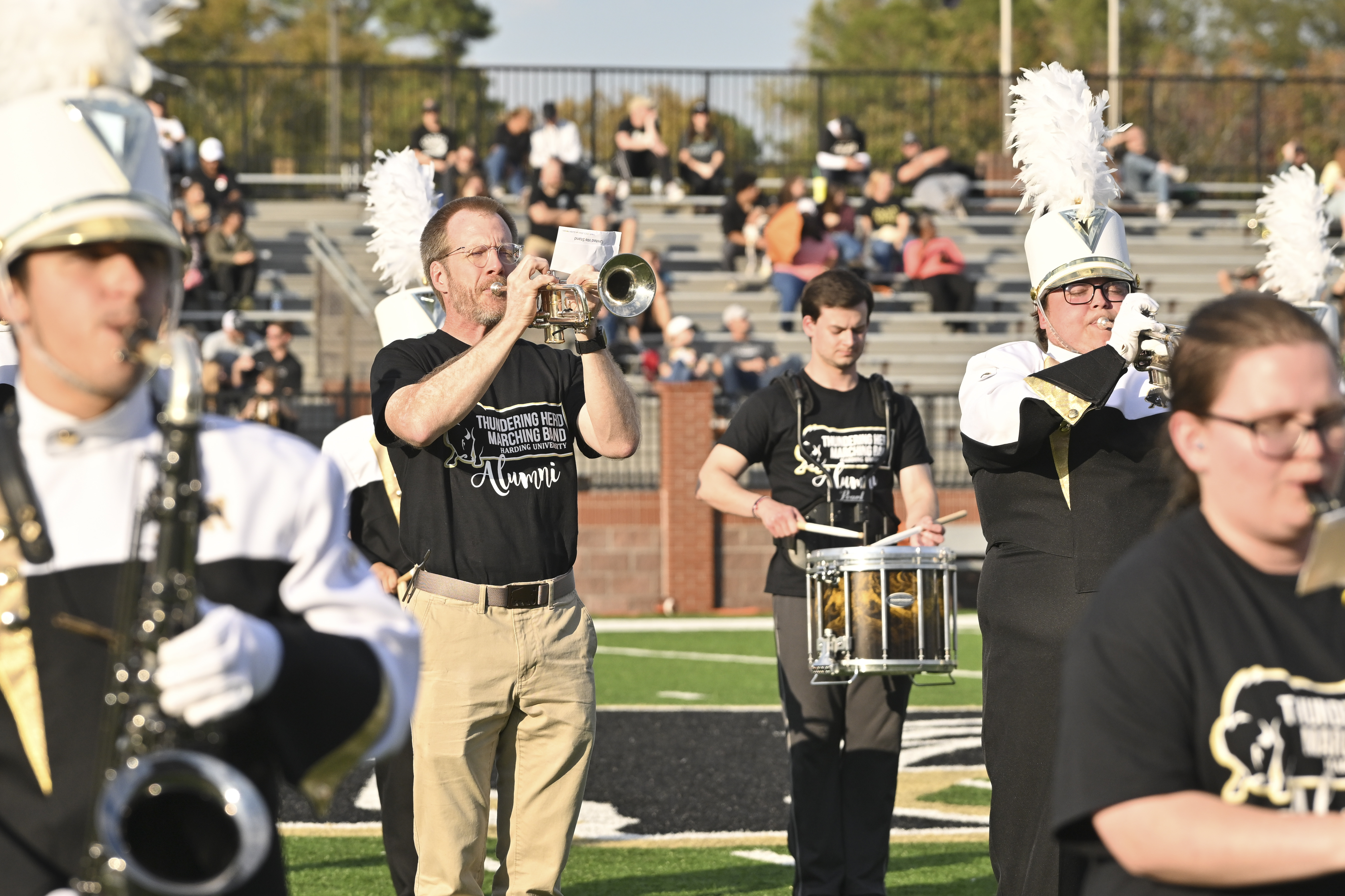 Marching band with band alumni playing on the Harding football field with spectators watching.