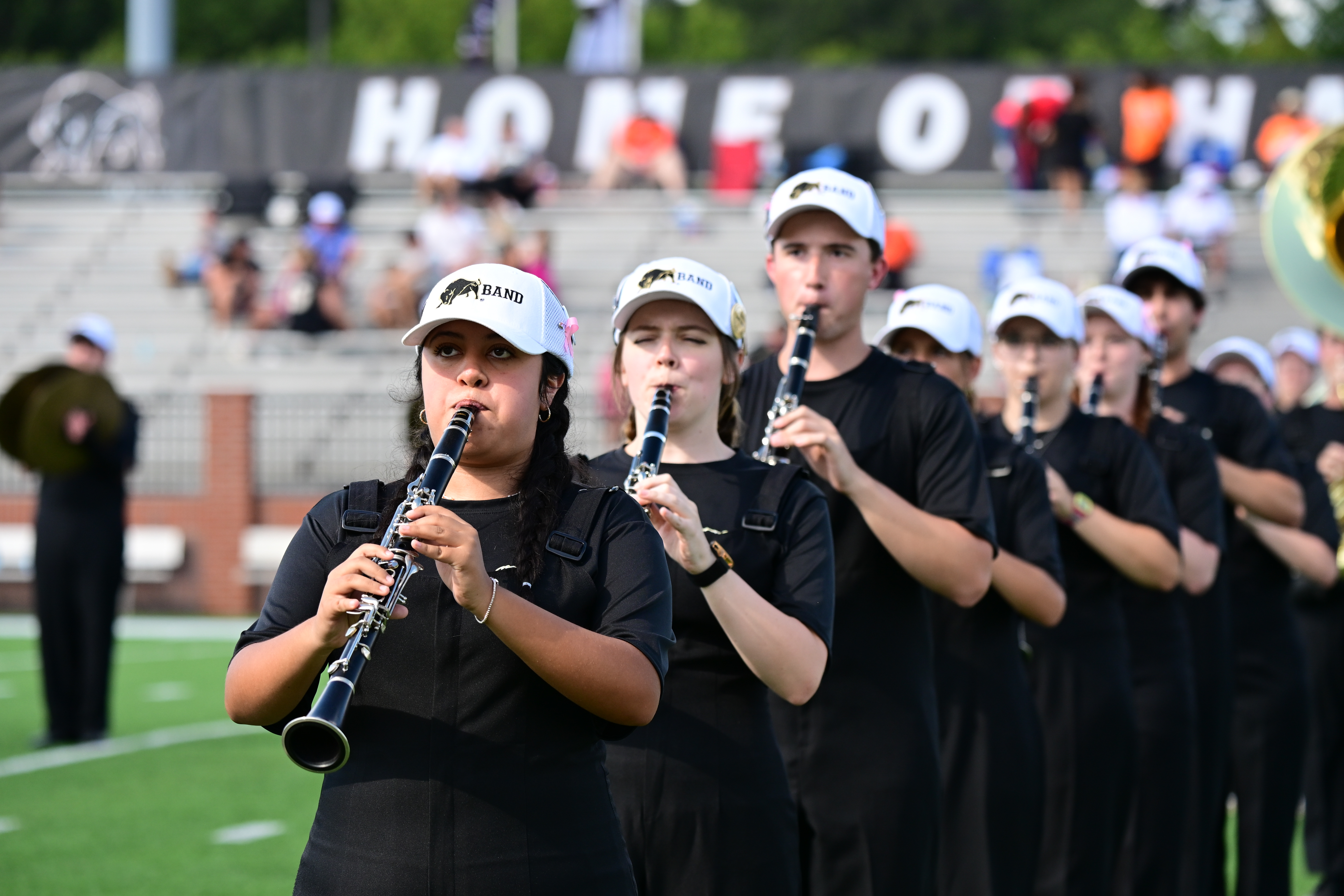 Marching band in a line formation dressed in black wearing white caps on Harding's football field with spectators watching.