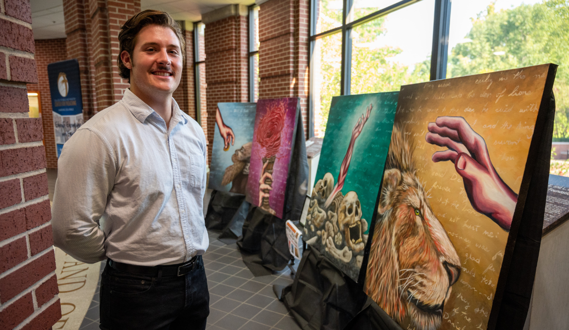 Nathan Mayes, a Harding Student wearing gray collared shirt smiles standing next to his winning pieces of art.