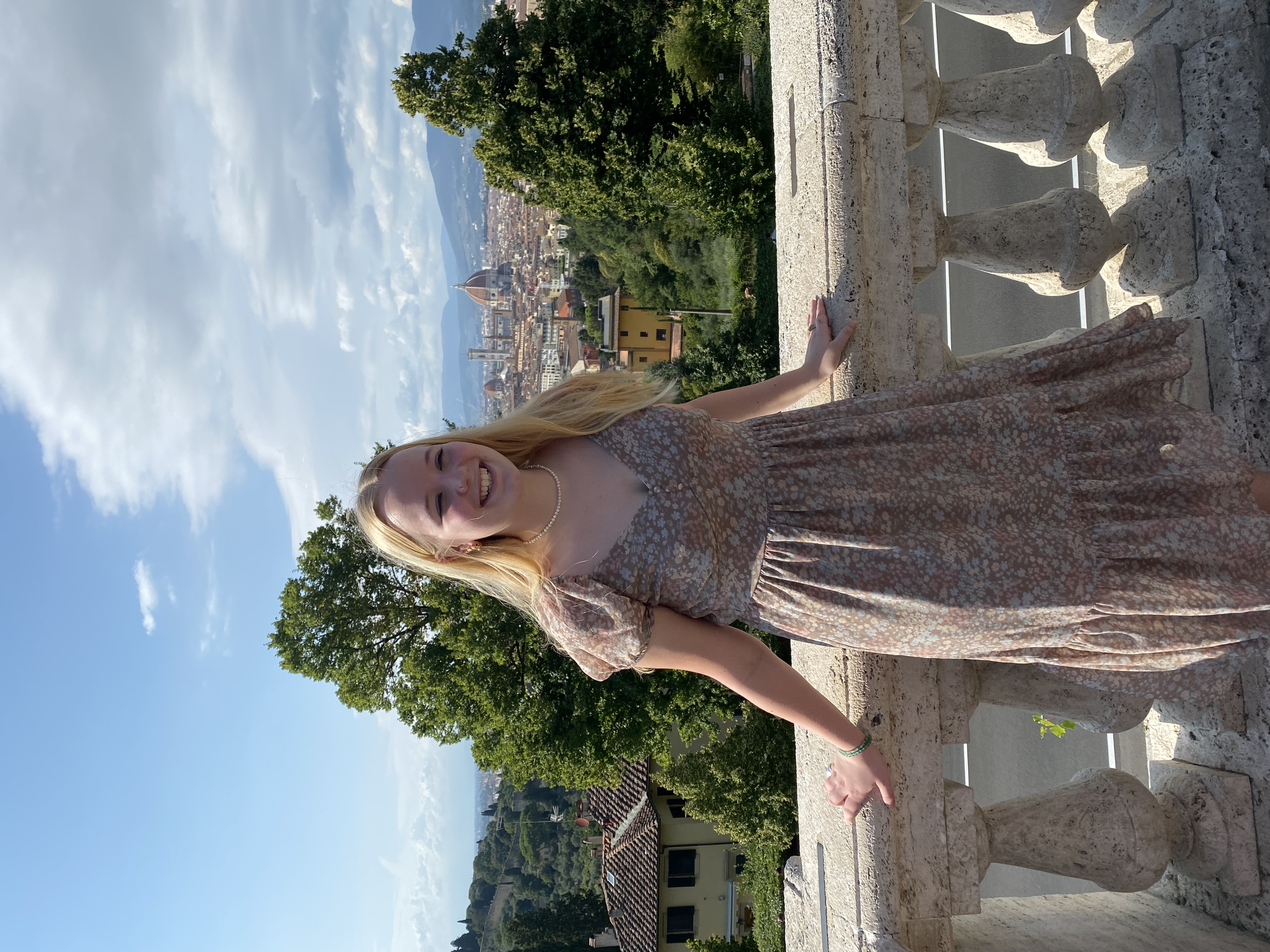 Maggie Samples smiles in a tan dress on a balcony with the sky behind her