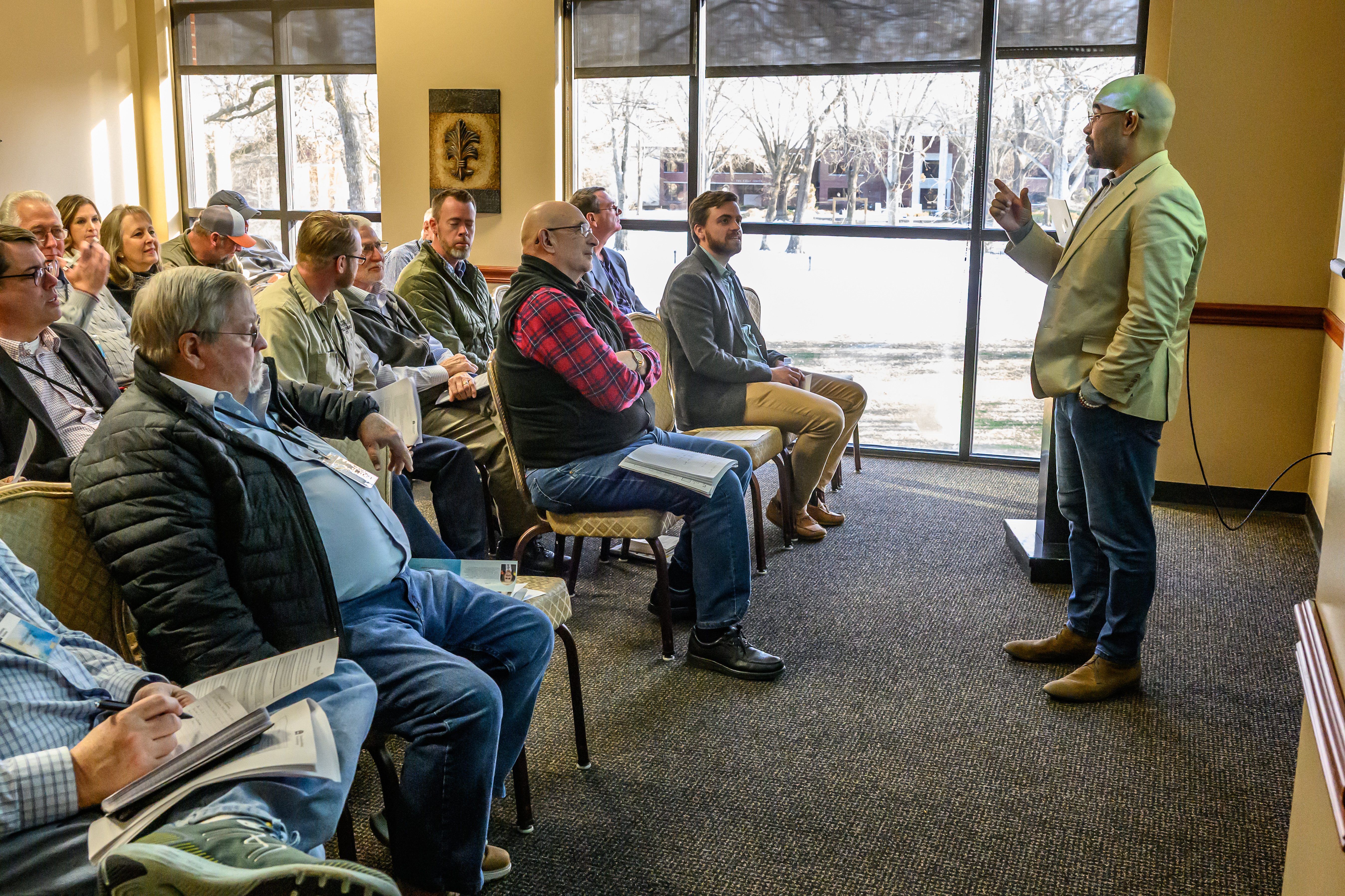 sMan in a sportscoat speaking in front of others in front of a large window overlooking trees