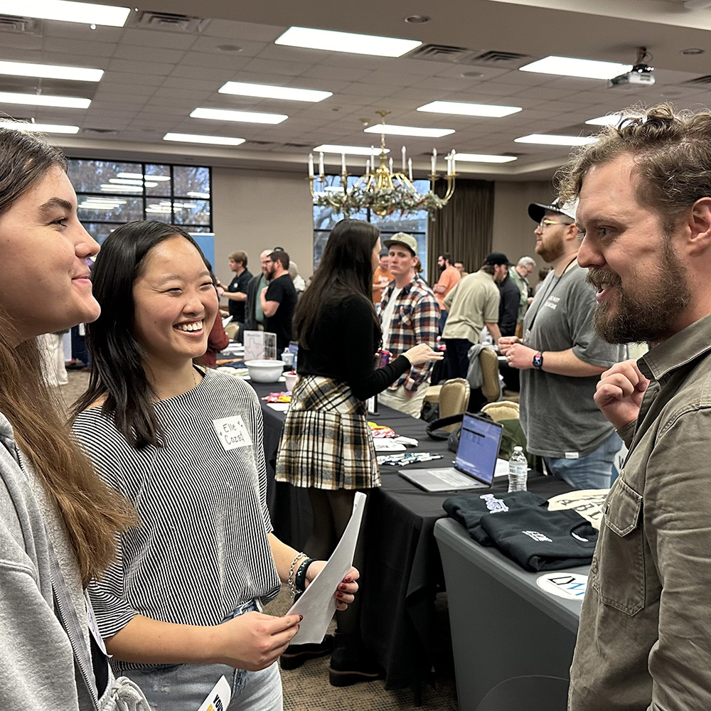 Two prospective youth interns talk with a man at a booth, with other booths and people behind them at the Youth Intern Fair.