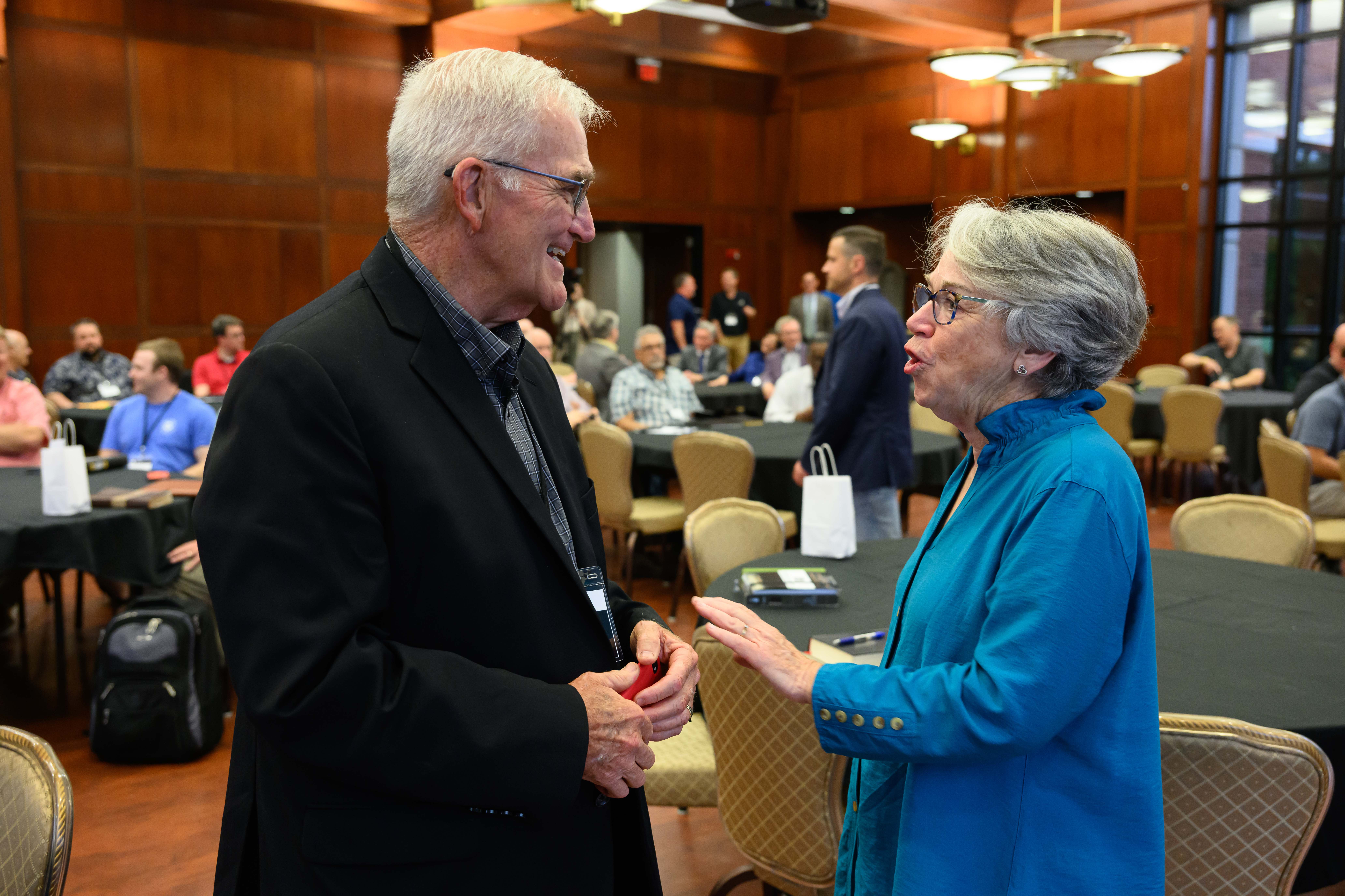 Dr. Phil Thompson talking to a woman with people seated behind them at round tables in Cone Chapel.