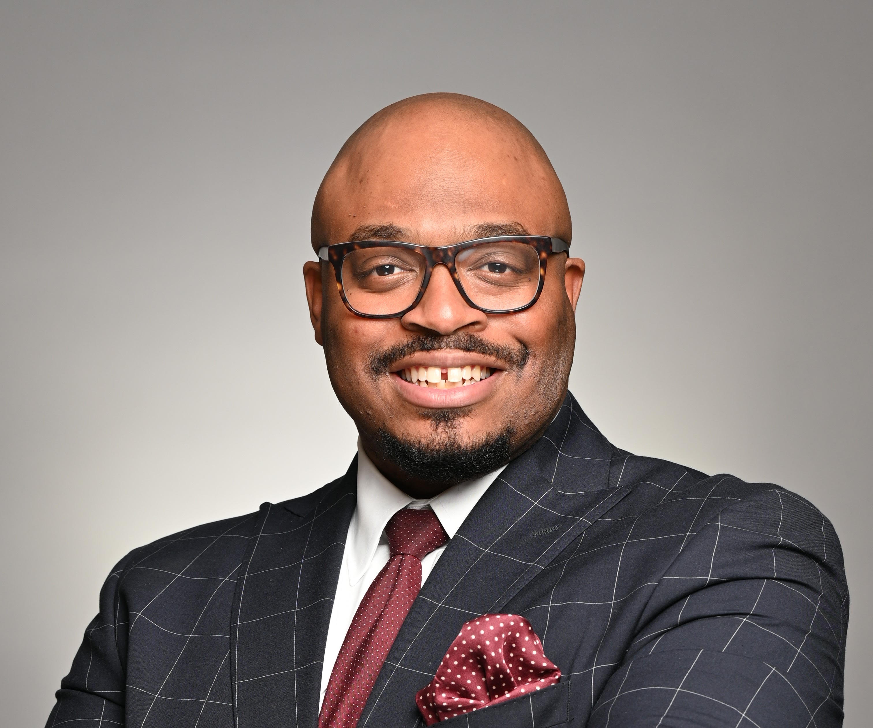 B.Chris Simpson smiles in glasses and a gray windowpane suit with burgundy tie and pocket square