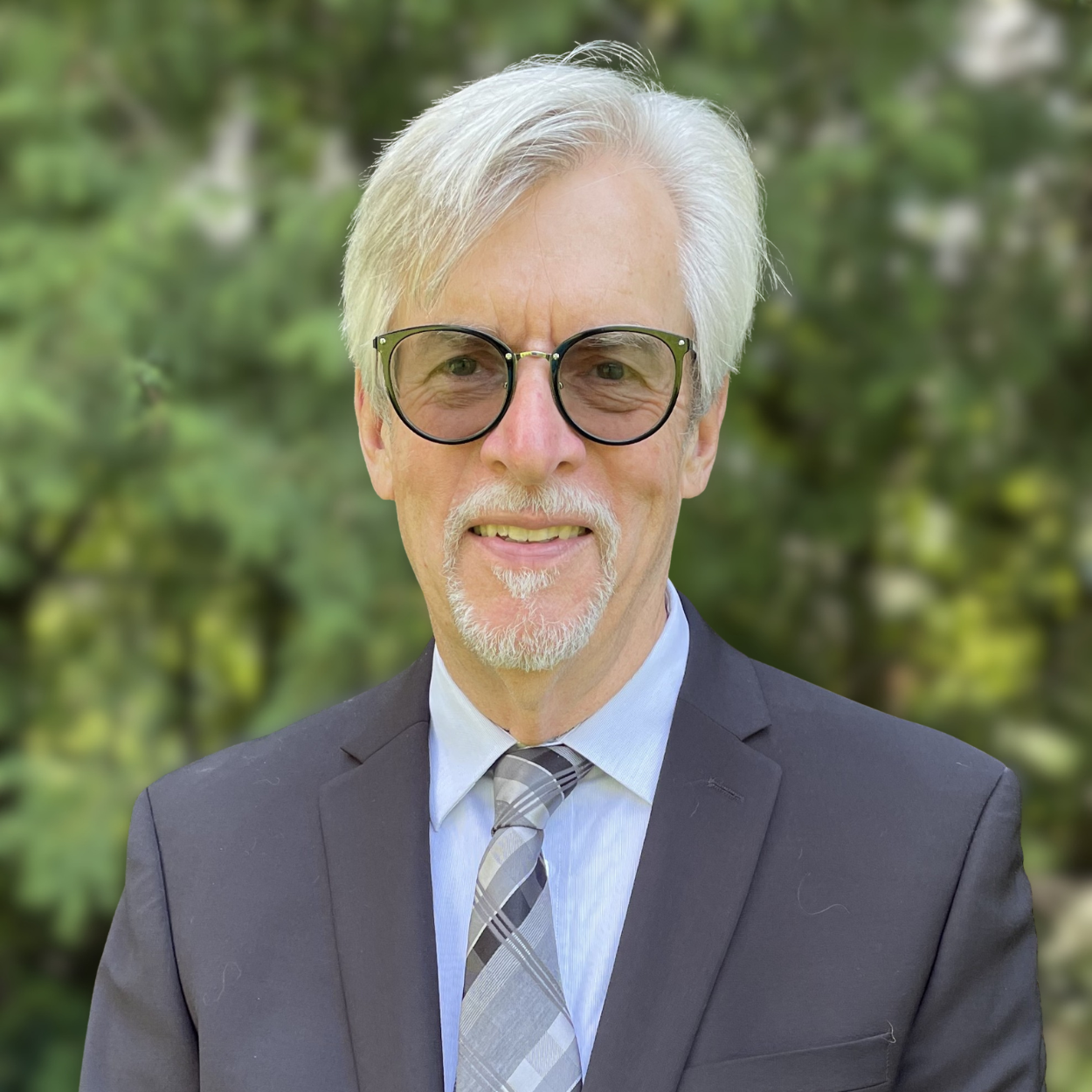 Dr. Paul Scott Wilson smiles in a suit and glasses outside infront of greenery