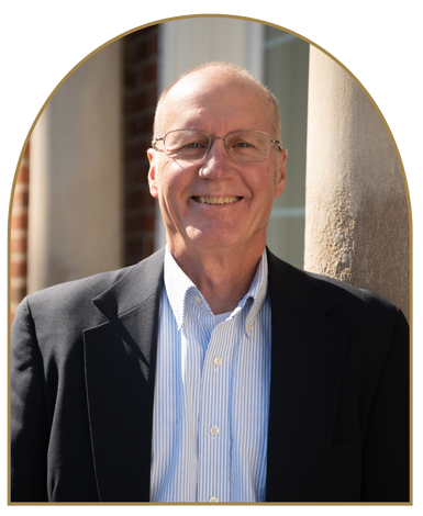 Portrait of Dr. Dave L. Bland smiling, wearing glasses, a dark blazer, and striped shirt by a brick building with columns.