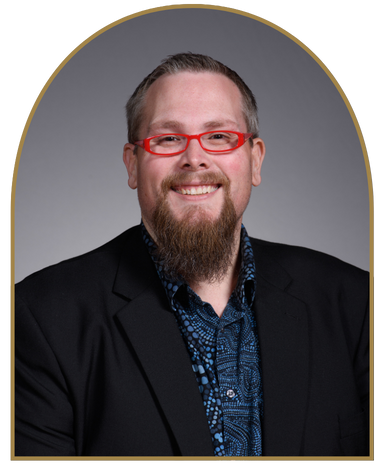 Portrait of Dr. Devin Swindle smiling wearing a black blazer with a blue collard shirt and red glasses on a gray background.