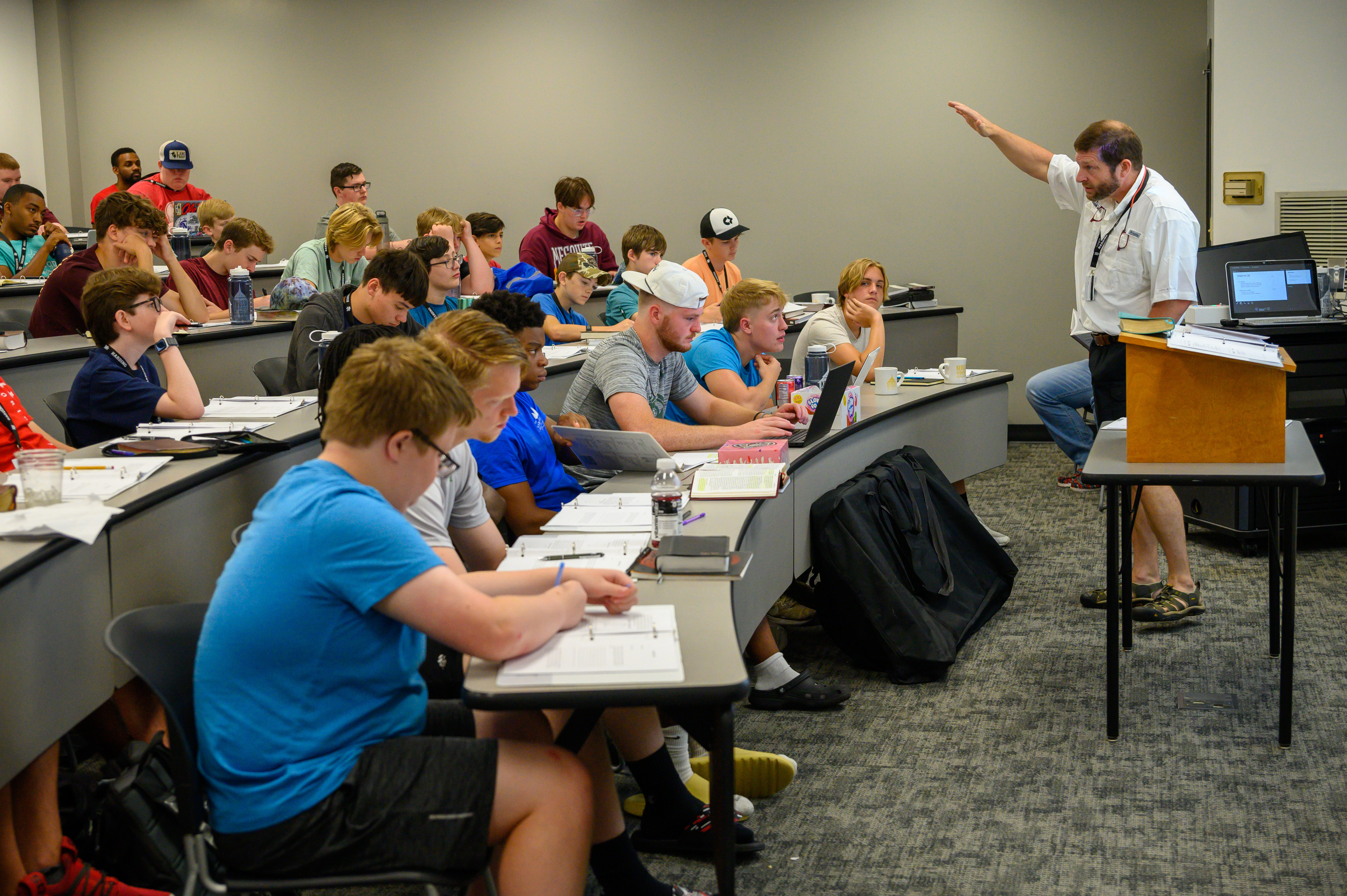 Man stands next to a table, raising his hand while presenting a slide presentation to a classroom full of people.