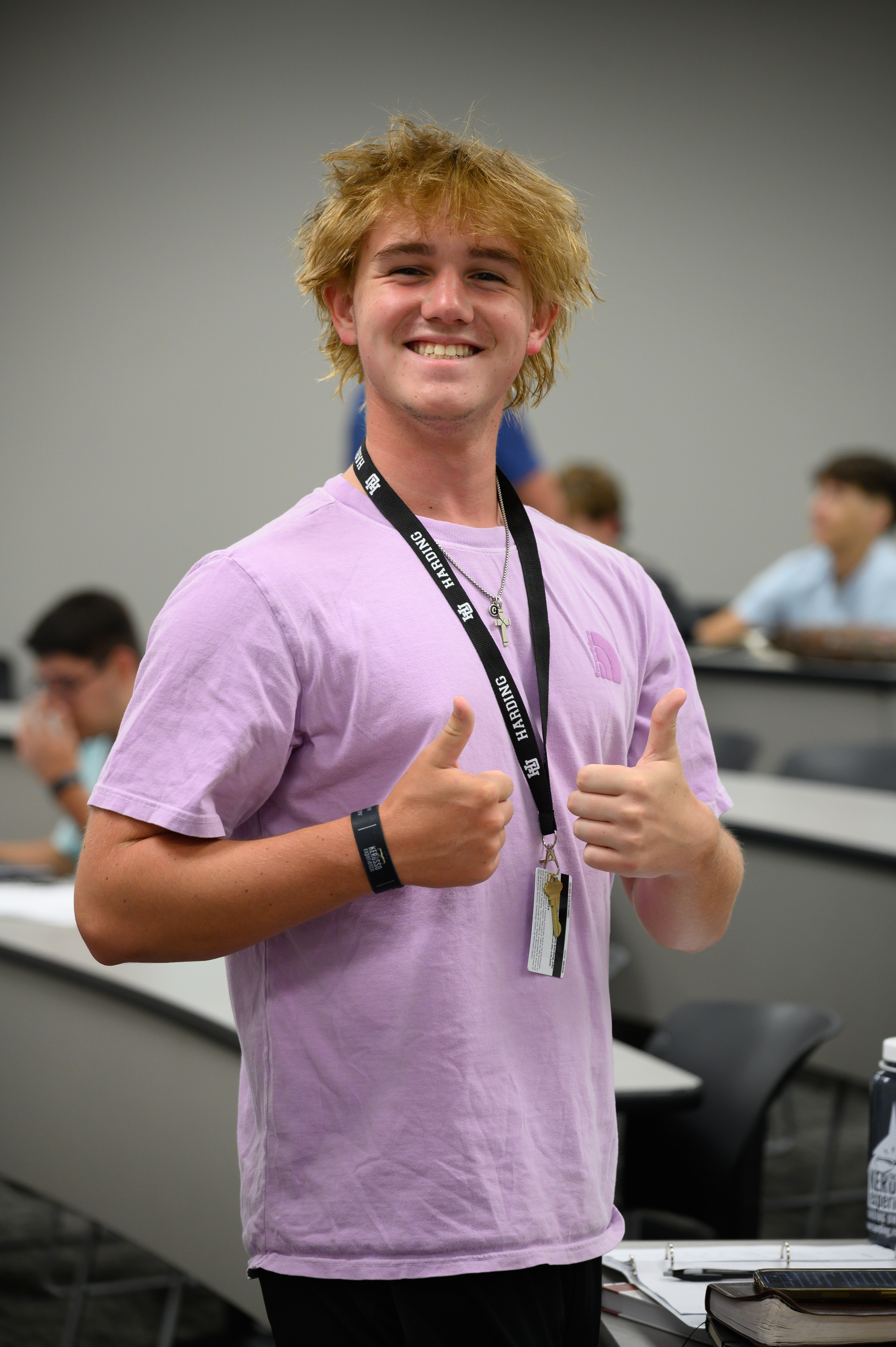 Student with blonde hair in a classroom, wearing a lilac shirt and Harding lanyard, smiles and gives two thumbs up.