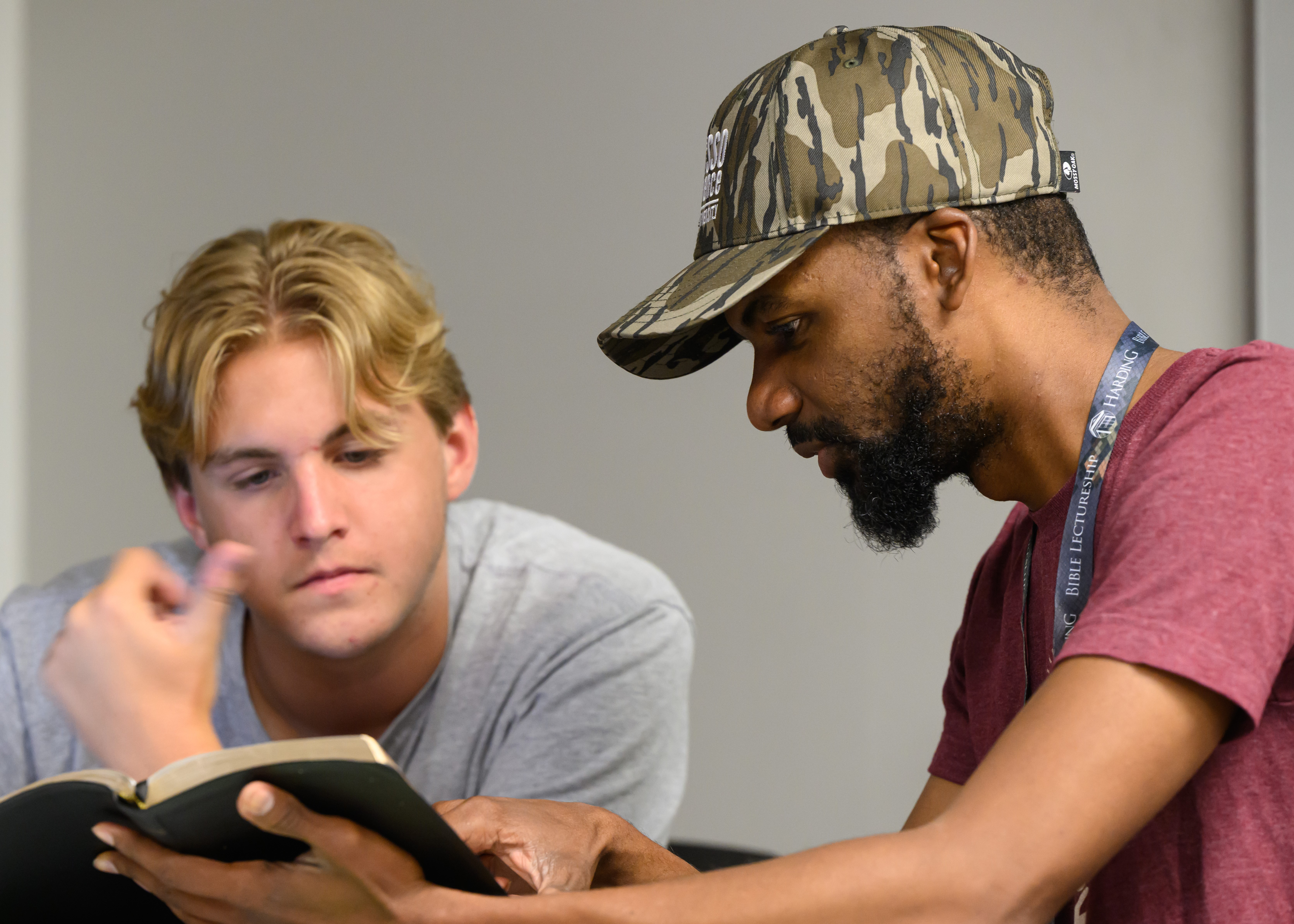 Man with dark hair and camouflage hat points to a Bible while sitting at a desk next to a boy with blond hair looking at the Bible.