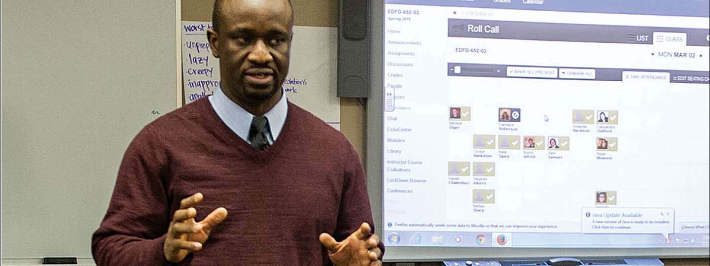 A man wearing a maroon sweater, blue shirt, and tie teaching while standing in front of a class with a projector.