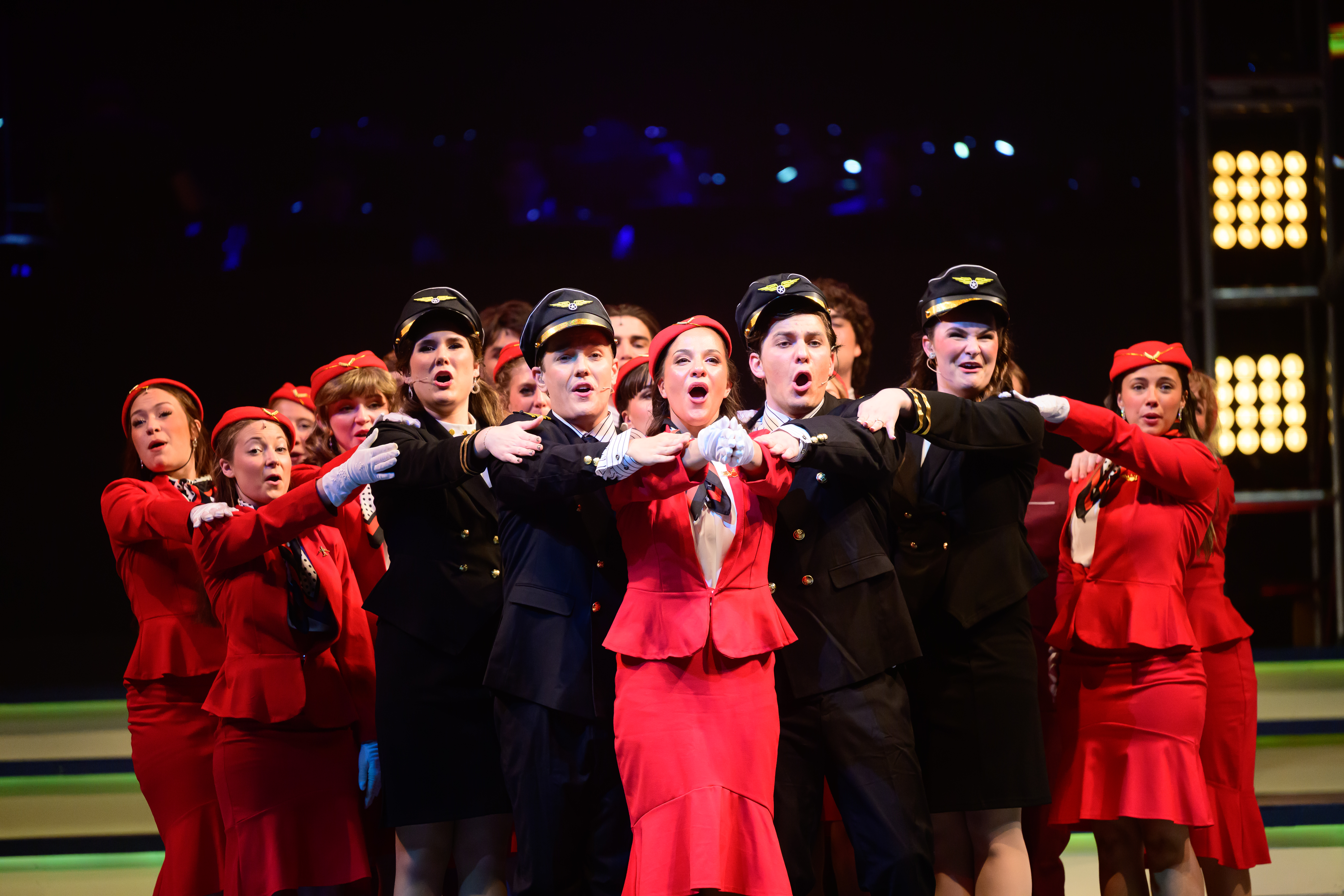 Members of the Spring Sing 2026 Ensemble dressed in flight attendant attire with arms outstretched to make the formation of an airplane.