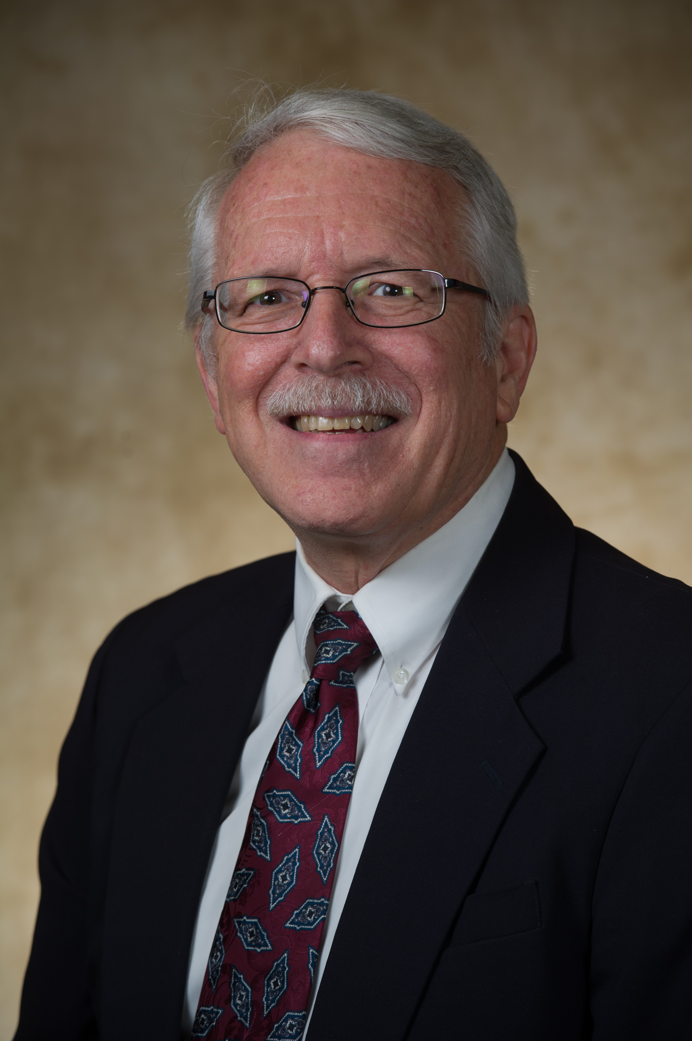 Allen Black, a man in glasses, dark suit, white dress shirt, and patterned tie against a soft beige background.