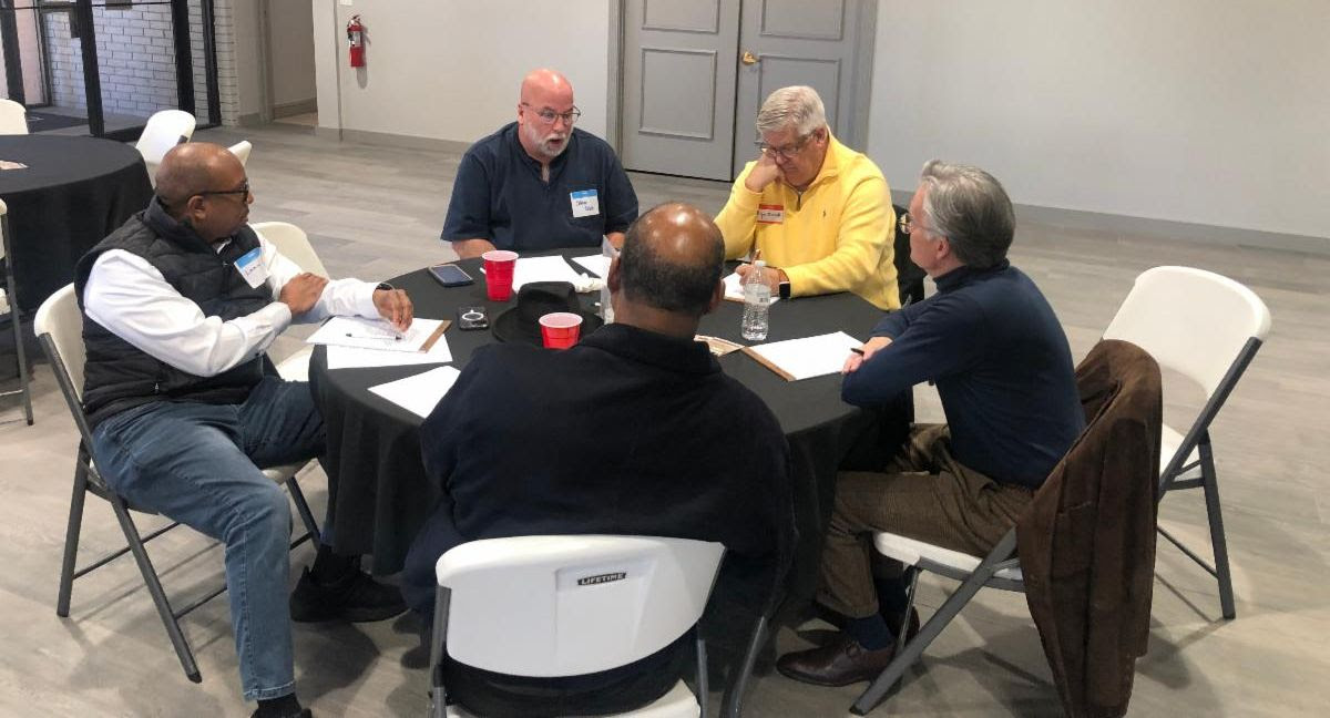 A group of men gathered around a table talking to each other.