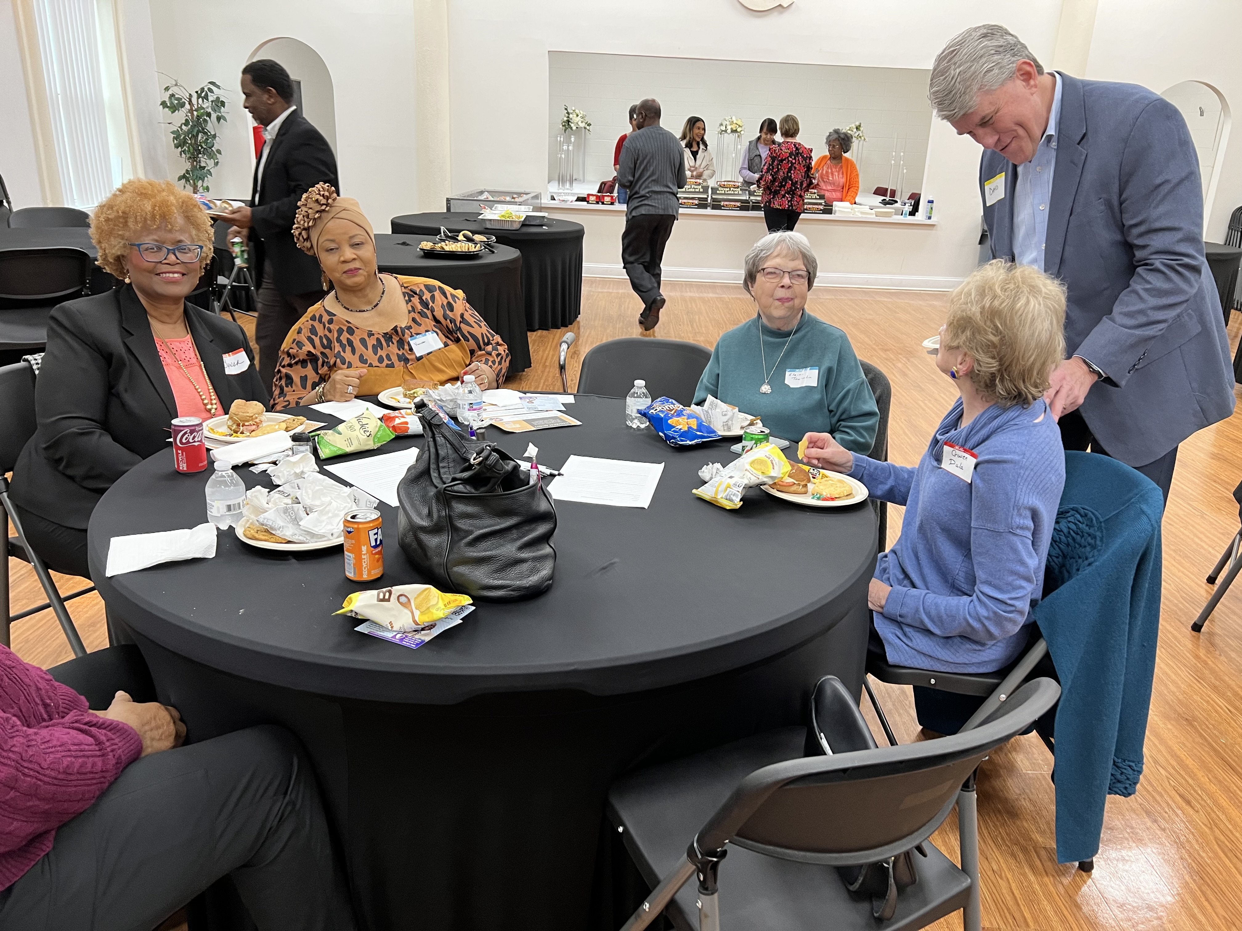 A group of people enjoying a meal at round tables, with food and drinks on the tables. Some are engaged in conversation.