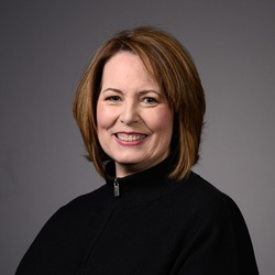 Chris Williams, smiling woman with brown hair wearing a black collared shirt against a gray background.