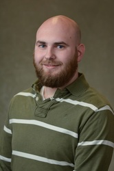 John Mark Warnick, smiling bald man with brown beard wearing a green collared shirt with white stripes on a gray background.