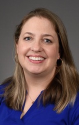 Lauren Boone, a woman smiling with brown hair, earrings, and a blue shirt against a gray backdrop.