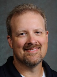 Dr. Patrick McGaha wearing a dark polo shirt smiling in front of a gray background.