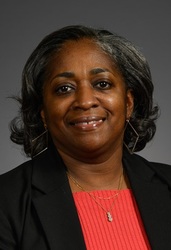 Tiffany Byers, a smiling woman with grayish-black hair, wearing a black blazer over a red shirt against a gray backdrop.