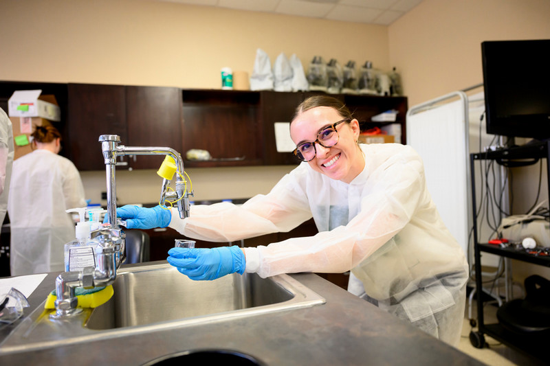 A pharmacy student smiles while wearing protective gear and works with a container of a lab sink